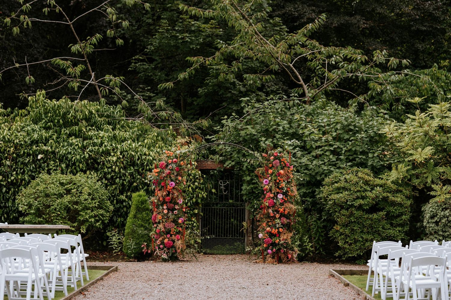 White wedding chairs arranged outdoors with lush greenery and red flowers in the background.