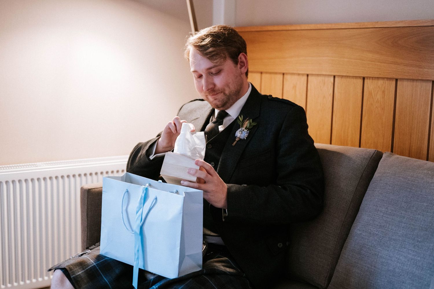 A person in a dark suit opens a light blue gift box while sitting on a couch.