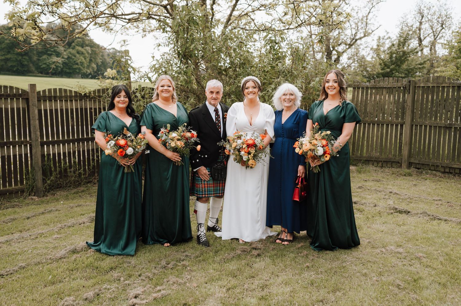 Wedding party in traditional Scottish attire and emerald dresses pose for formal photo.