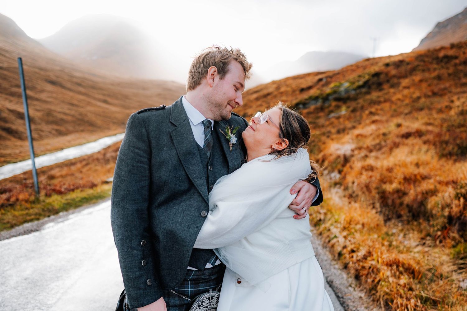 A romantic wedding couple embraces on a misty mountain road in the Scottish Highlands.
