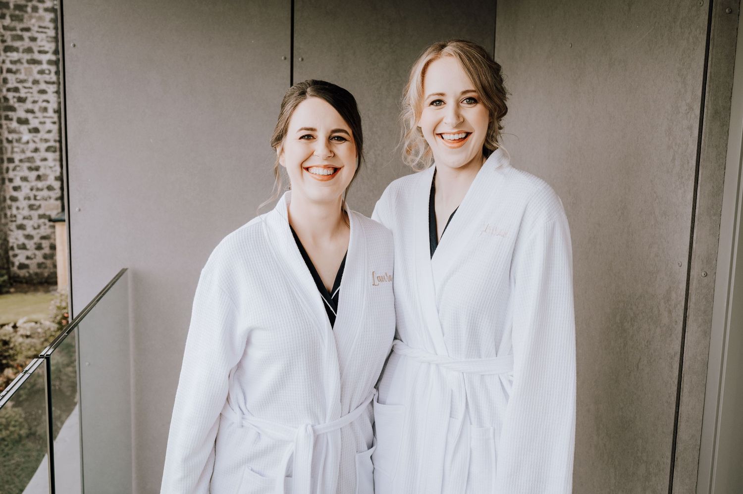 Two people in white robes smile while standing against a brick wall.