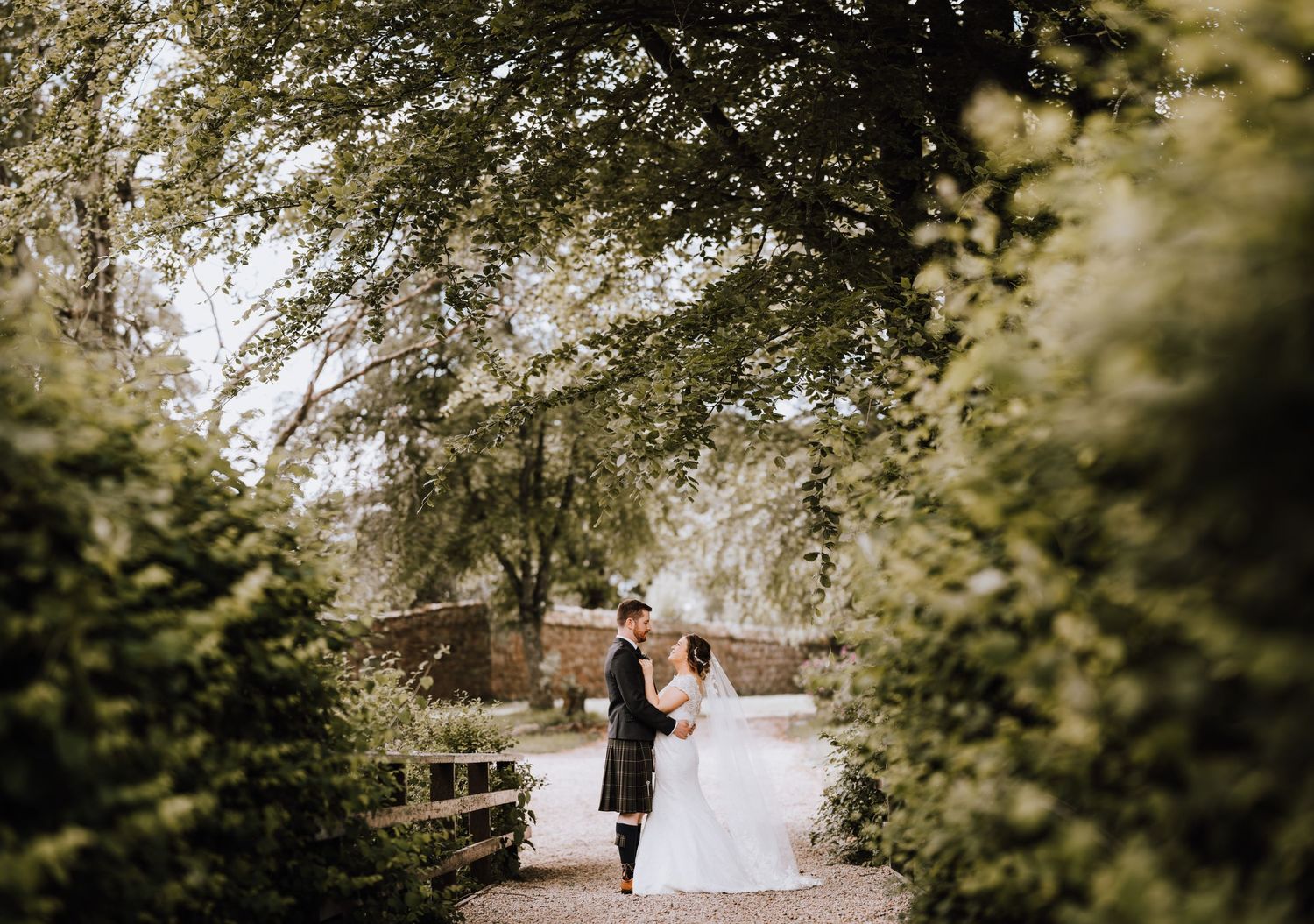 A scenic wedding photo framed by trees showing a couple in traditional Scottish attire on a garden path.