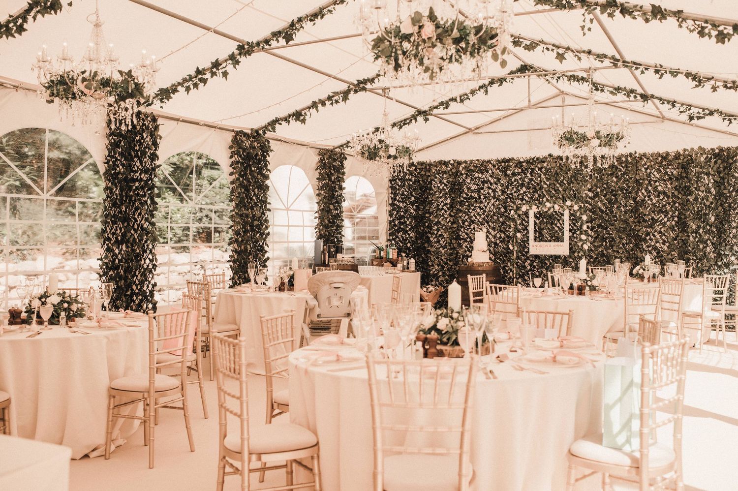 Elegant wedding reception tent decorated with greenery garlands and white tables with gold chiavari chairs.