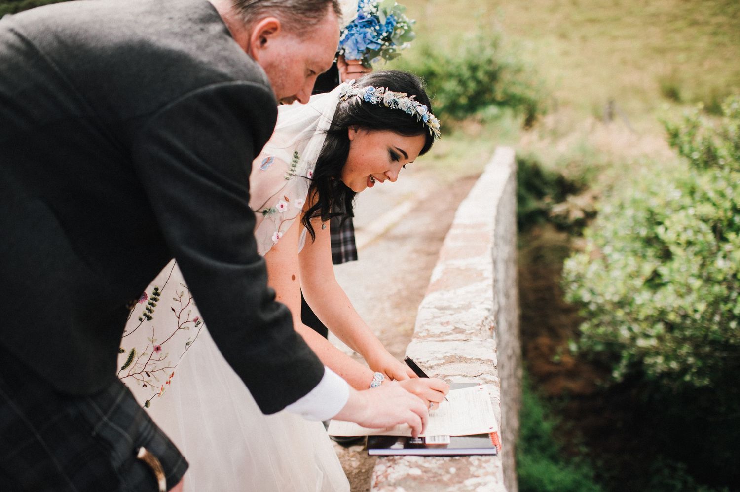 Wedding participants sign official documents on the stone wall of a bridge in the Scottish countryside.
