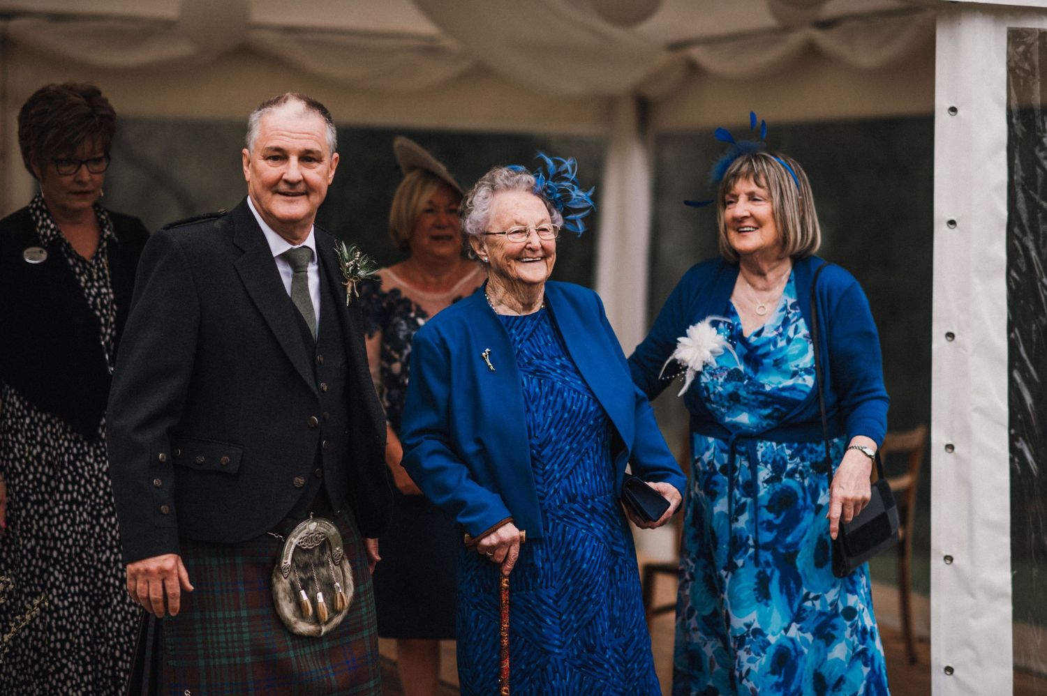Wedding guests in formal attire gather under white tent wearing traditional Scottish clothing.