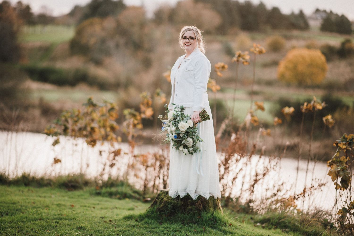 A bride in white dress holds bouquet while standing by autumn lakeside.