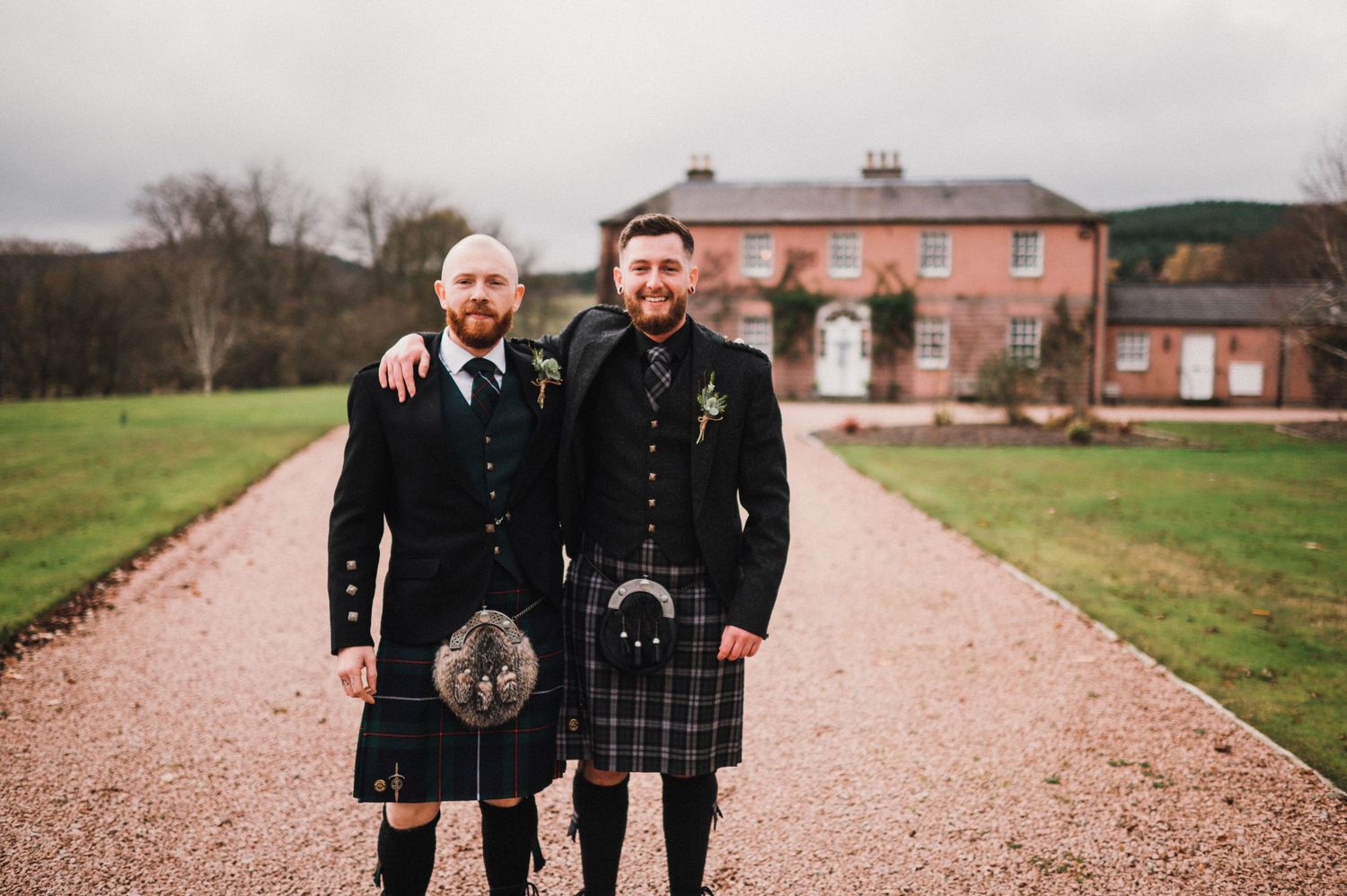 Two people wearing traditional Scottish kilts stand together on a gravel path in front of a brick manor house.