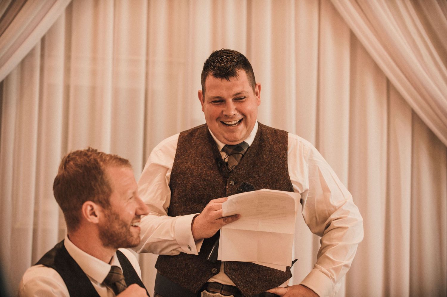Two guests sharing a humorous moment while reading papers at a wedding reception.