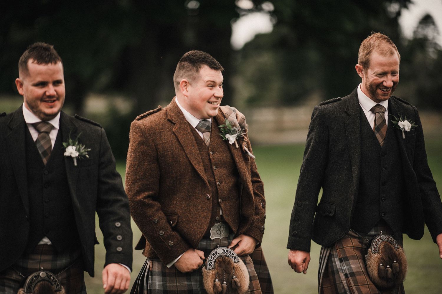 Three groomsmen in kilts and formal wear share a laugh during an outdoor event.
