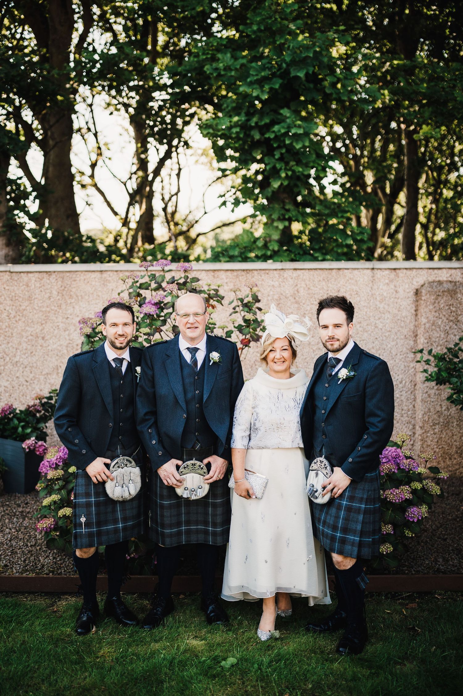 Wedding party in traditional Scottish formal wear poses against garden wall.