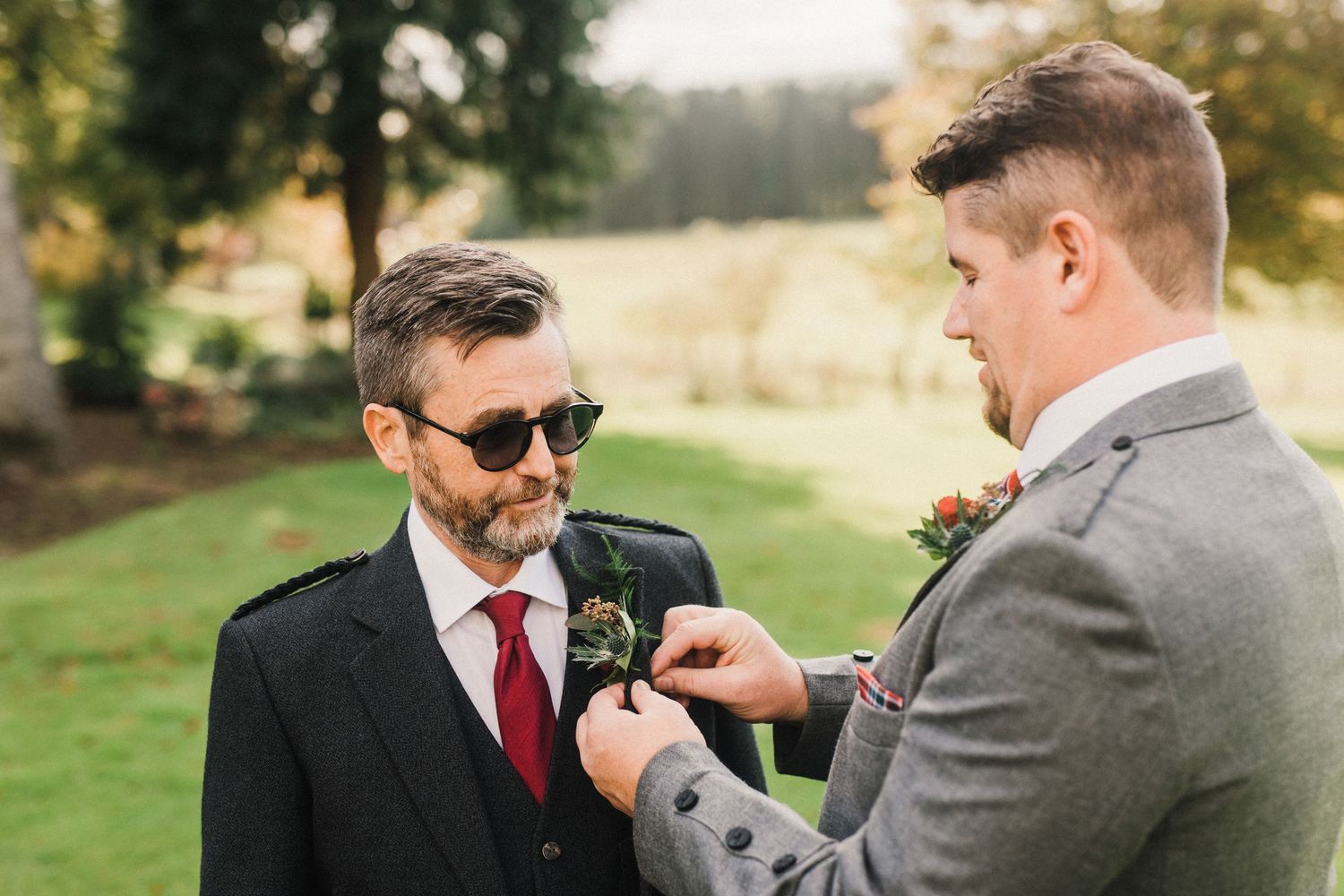 Two people in formal wear outdoors while one adjusts the other's boutonniere.