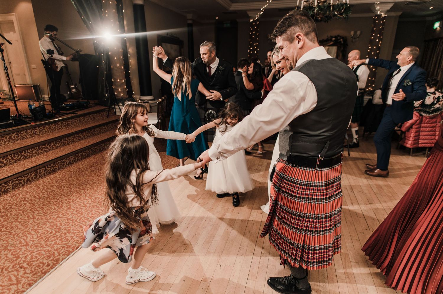 Wedding guests dancing together on a wooden floor with Scottish kilts and formal attire.