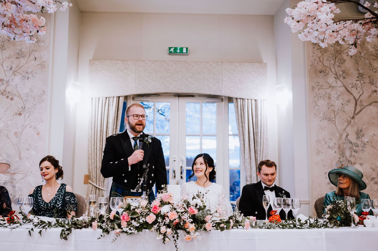 Wedding party seated at decorated head table with floral arrangements and Scottish attire.