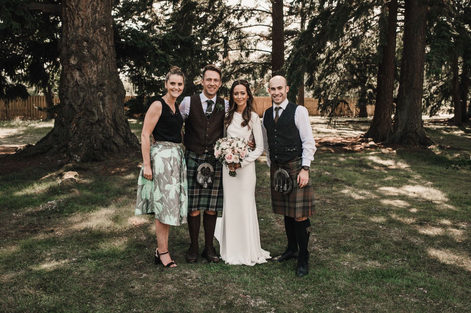 Wedding party in Scottish attire poses for formal photo in forest setting.