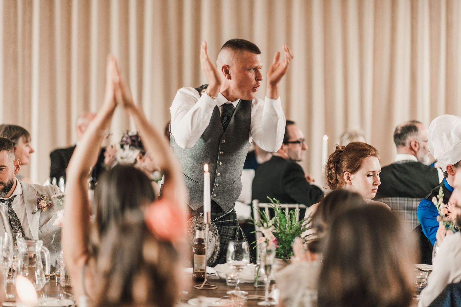 Reception guests raise their hands in celebration during a wedding speech at an indoor venue.