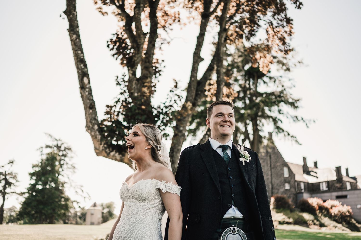 A couple in wedding attire poses together outdoors with trees and architecture in the background.