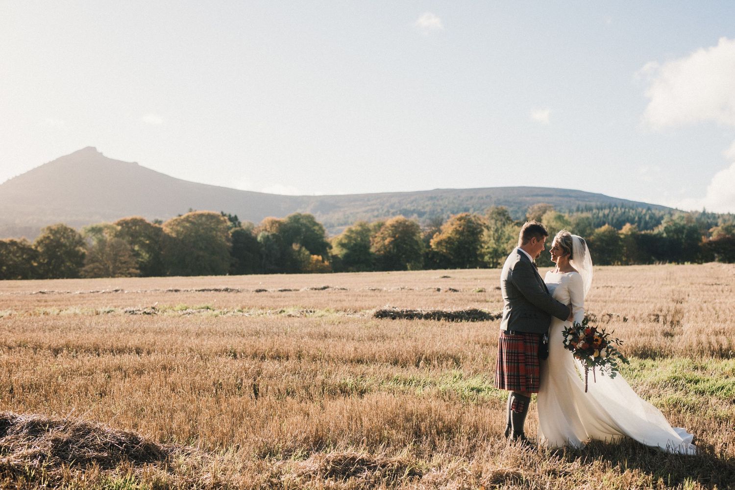Newlyweds embrace in golden field with mountain vista and autumn trees in background.