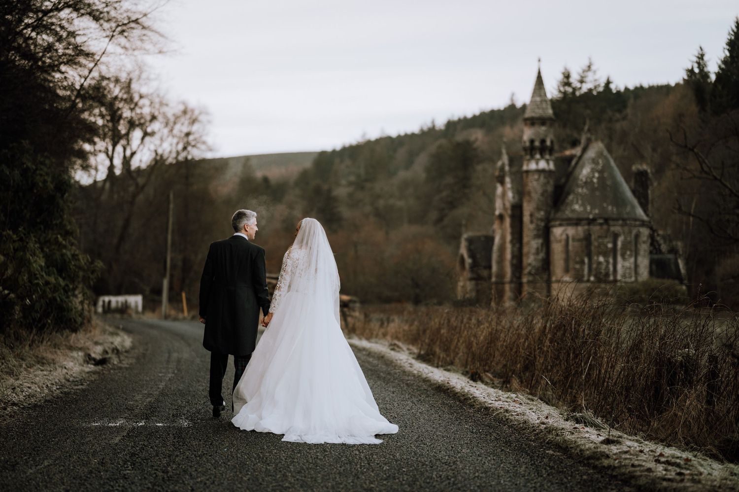 Wedding couple walking along a scenic country road with a gothic chapel in the distance.