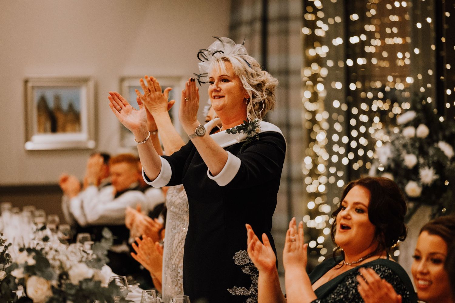 Wedding guests clapping and celebrating during reception with twinkle lights and festive atmosphere.