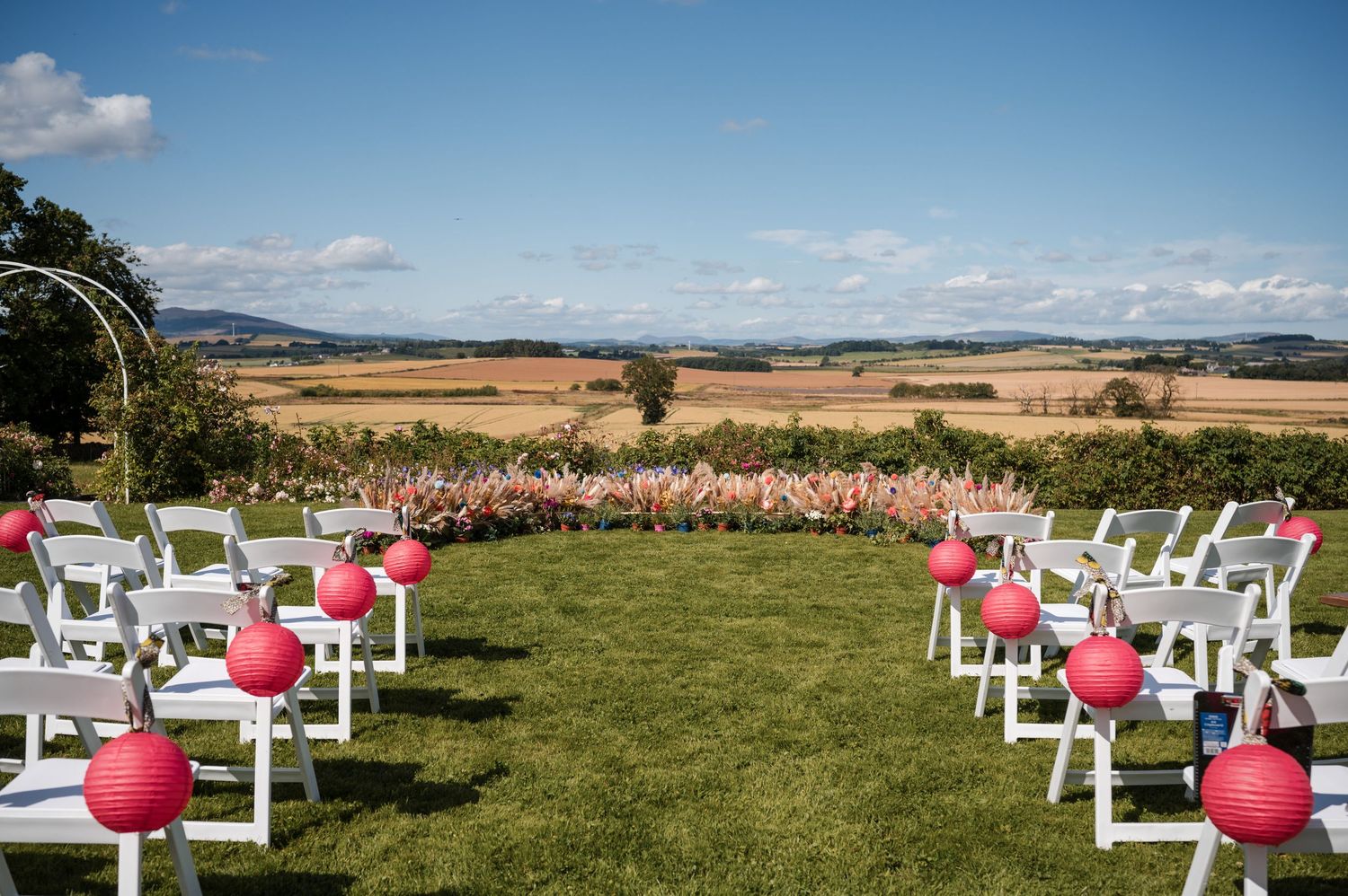 White chairs arranged outdoors for a wedding ceremony with pink balloons under a bright blue sky.