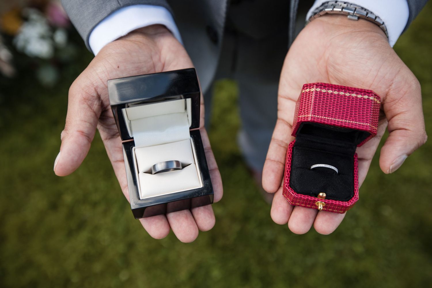 Two hands holding open ring boxes displaying wedding rings against a grassy background.