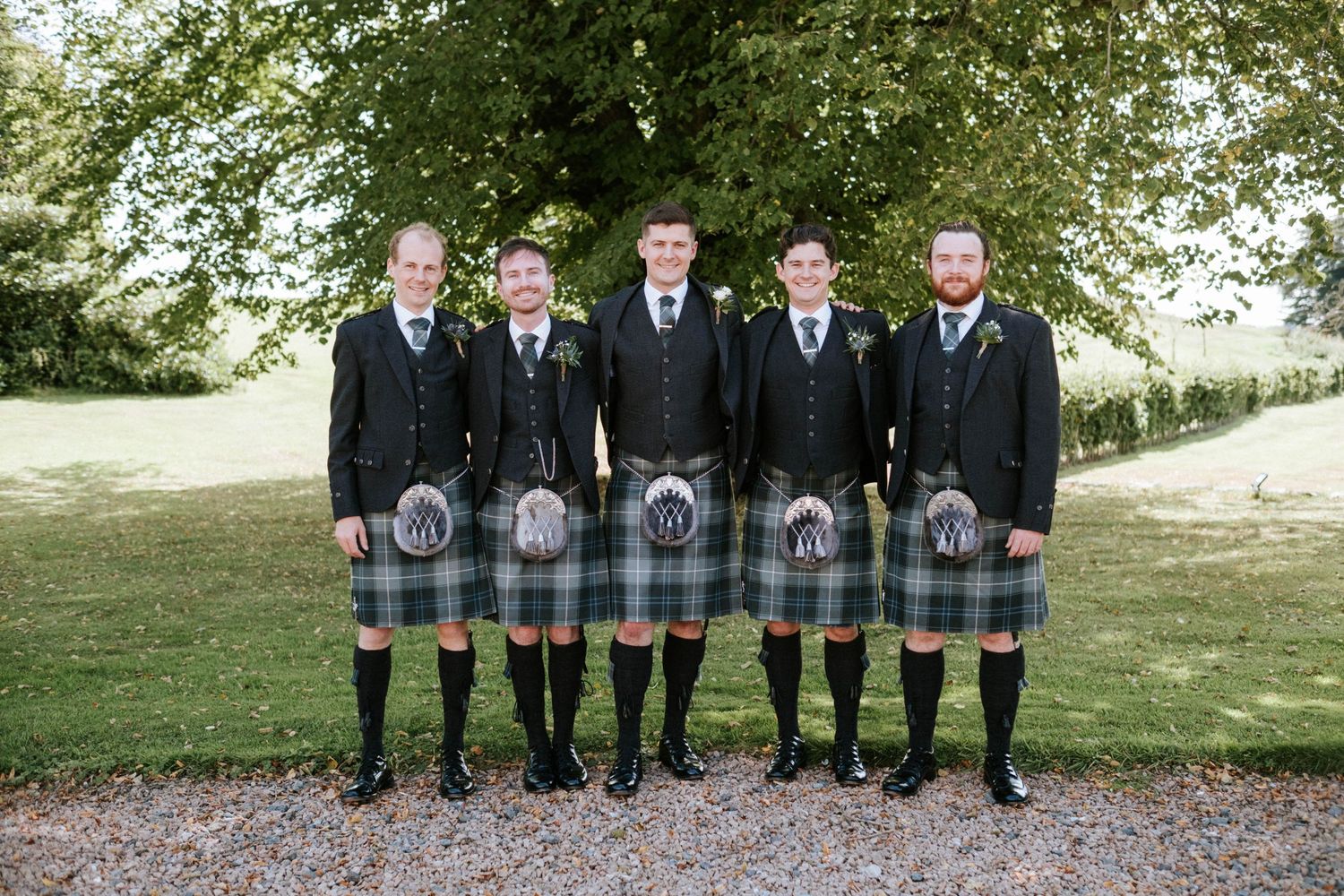 Groomsmen lined up wearing matching kilts and formal jackets under green trees.