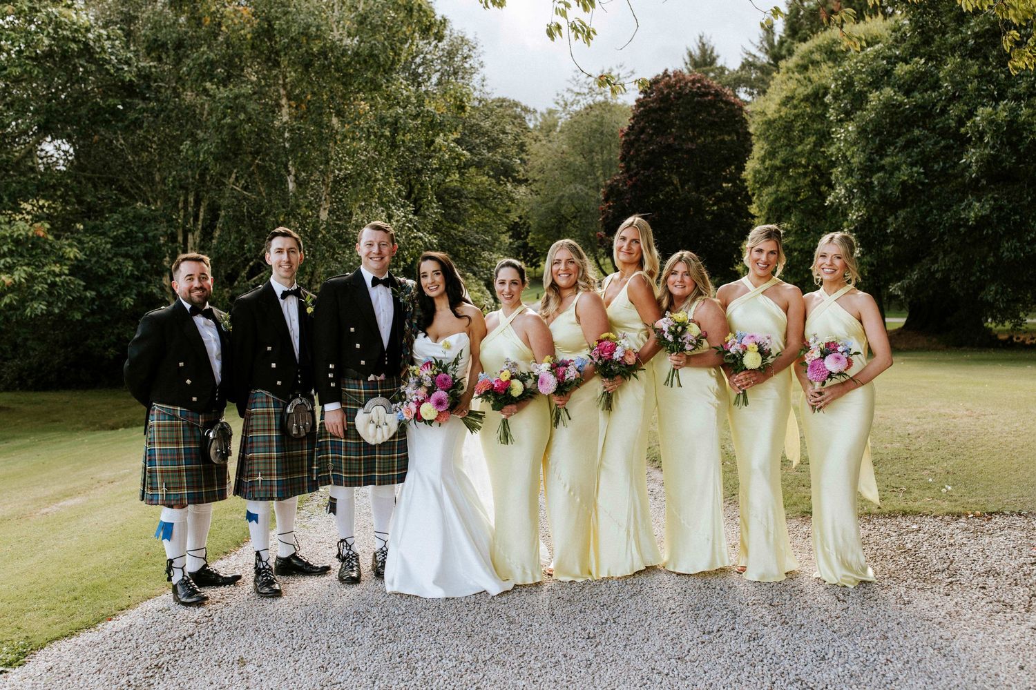 Wedding party poses together on gravel path with bridesmaids in cream dresses and groomsmen in kilts.