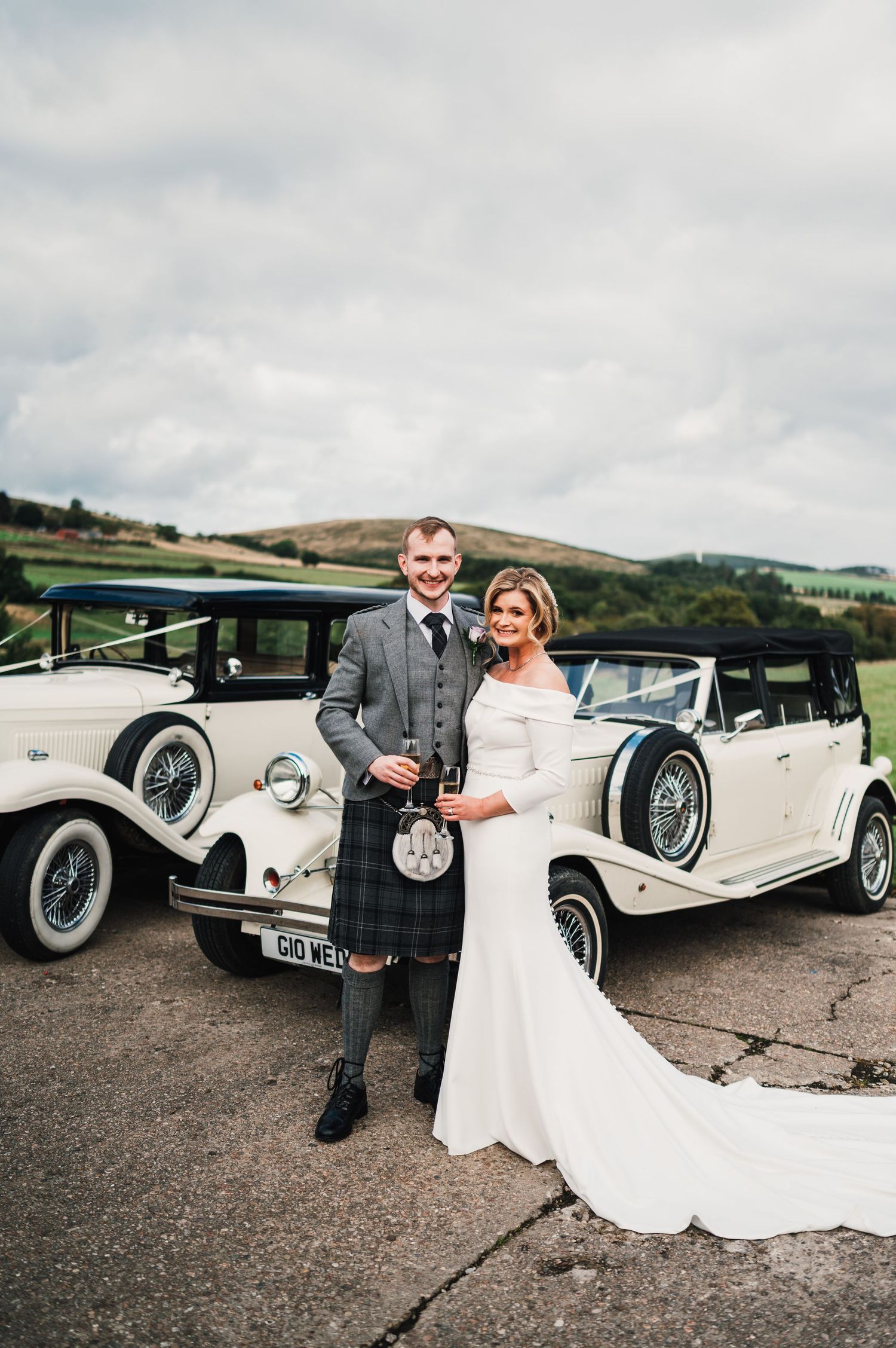 Classic white vintage wedding cars lined up against cloudy Scottish countryside backdrop.