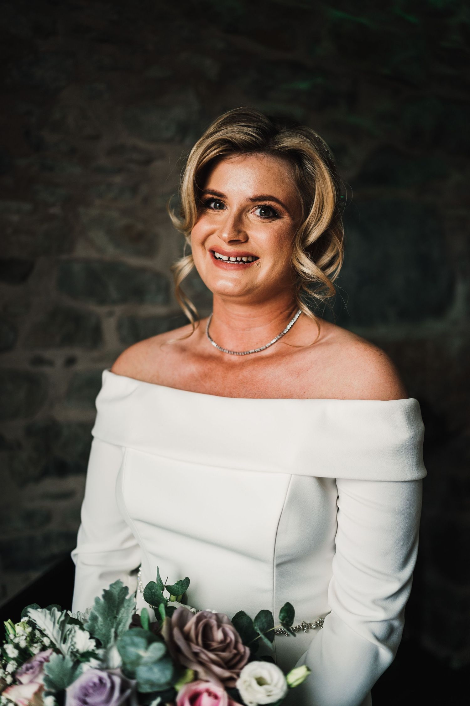 Close-up portrait of a bride in an elegant off-shoulder white wedding dress holding a purple and white bouquet.