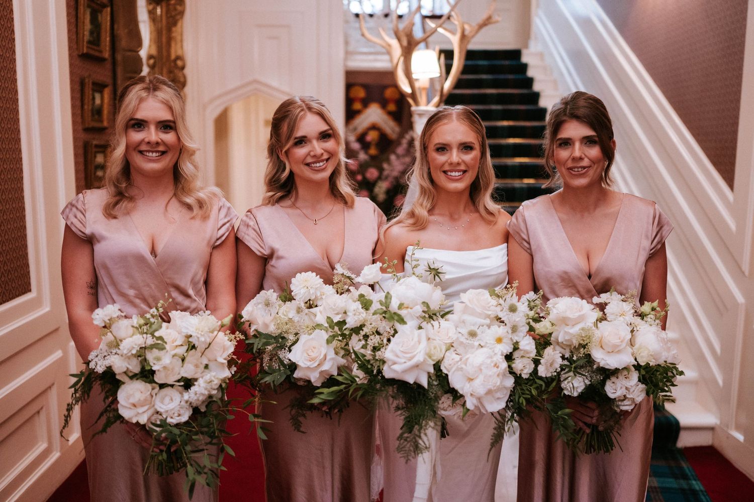 Bridesmaids in blush pink dresses hold elegant white floral bouquets while posing together.