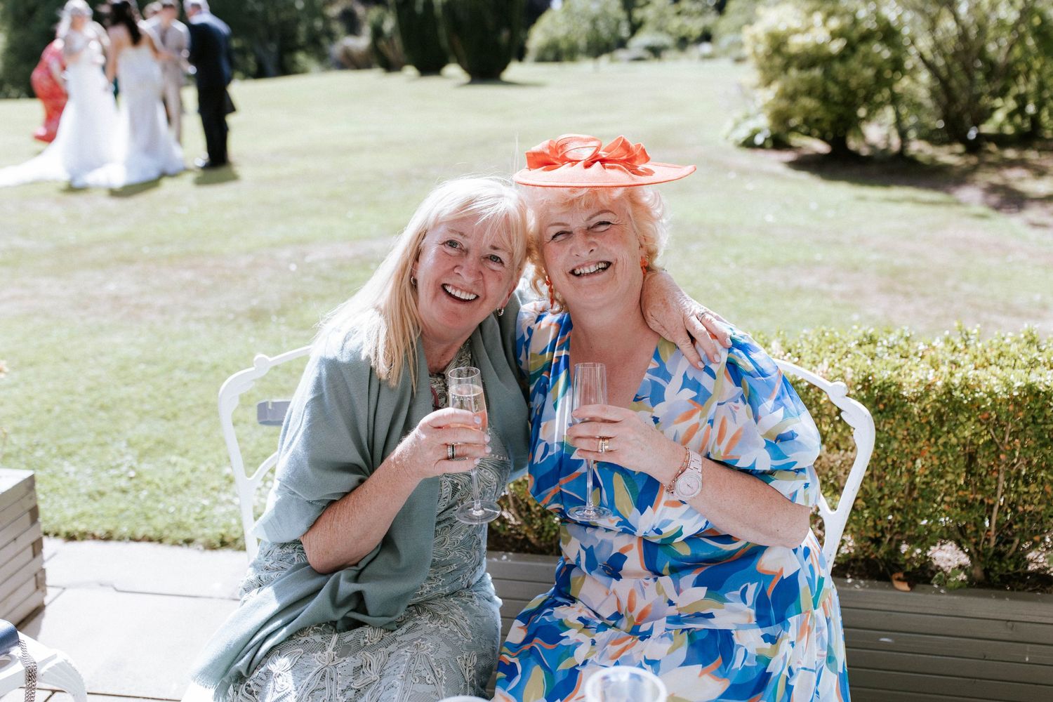 Two wedding guests sharing laughter while enjoying drinks in garden setting.