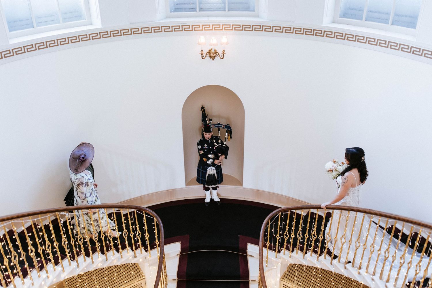 Three guests stand on an elegant curved staircase with ornate gold railings in a white-walled venue.