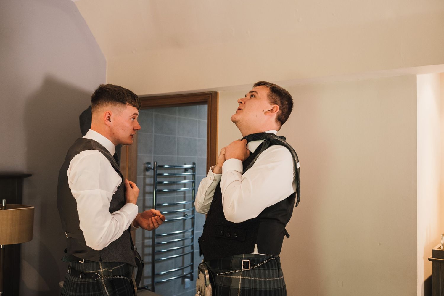 Two men in matching kilts and vests prepare for a formal Scottish event indoors.