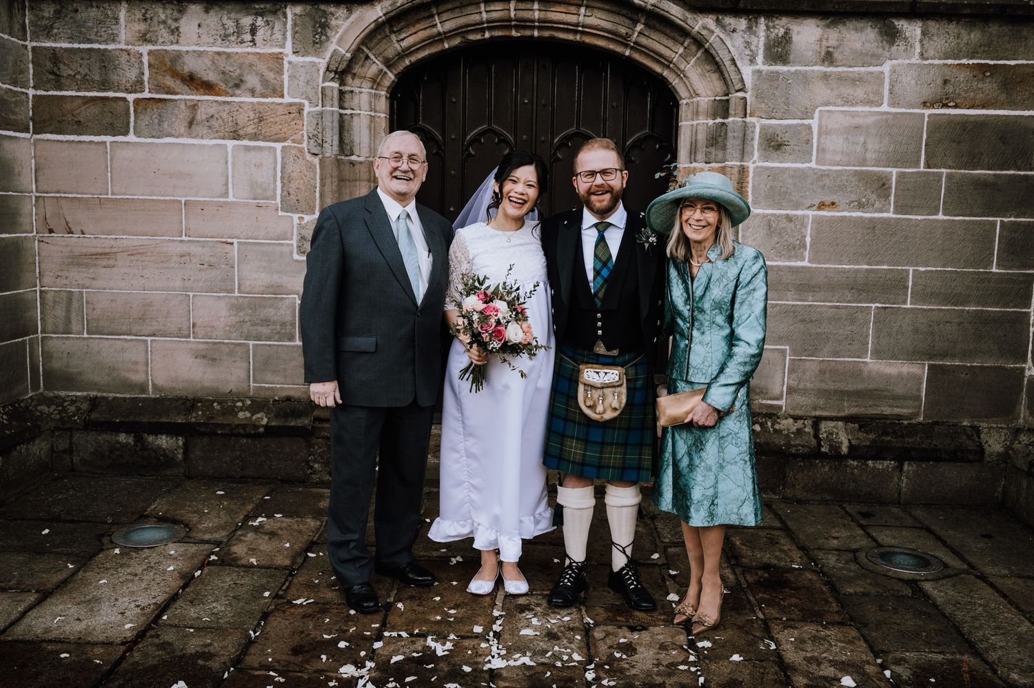 Small wedding group poses by stone church doorway with groom in traditional Scottish kilt.
