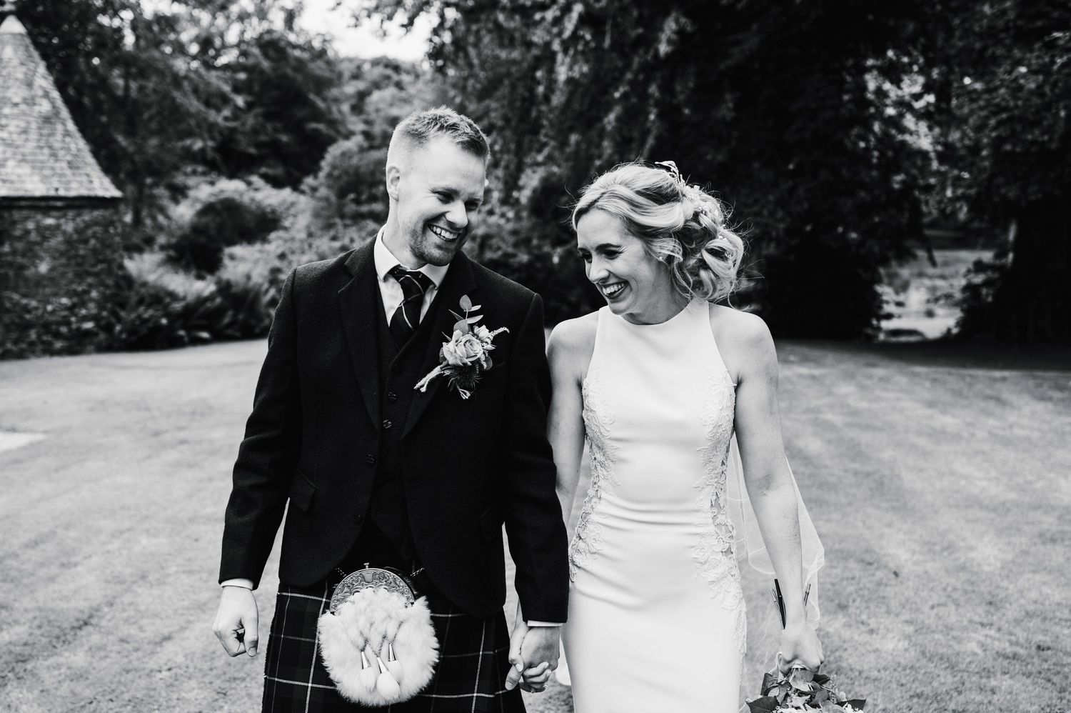 Black and white photo of a joyful couple walking outdoors, with groom in traditional Scottish attire.