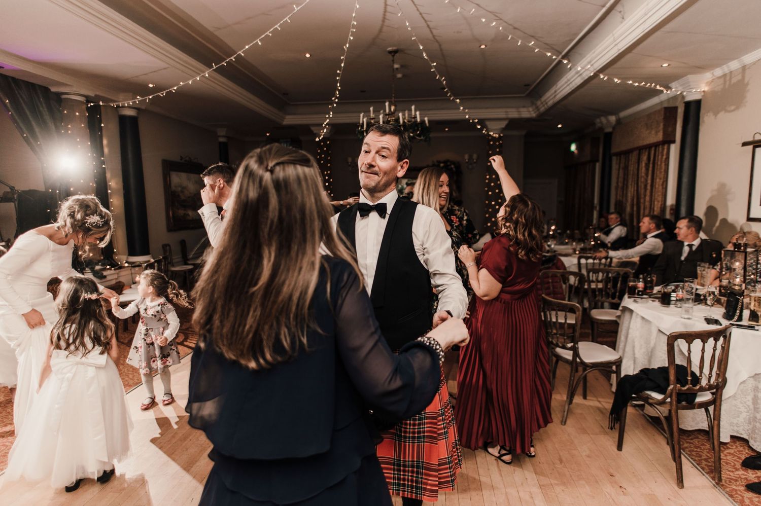 Wedding reception dance floor scene with guests celebrating under string lights and chandeliers.