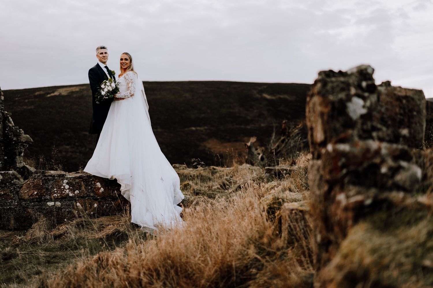 Newlyweds pose against dramatic landscape with ancient stone ruins in the foreground.
