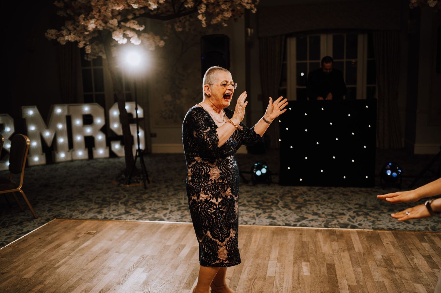 Person in sparkly dress dances enthusiastically at an evening reception with illuminated 'MRS' sign.