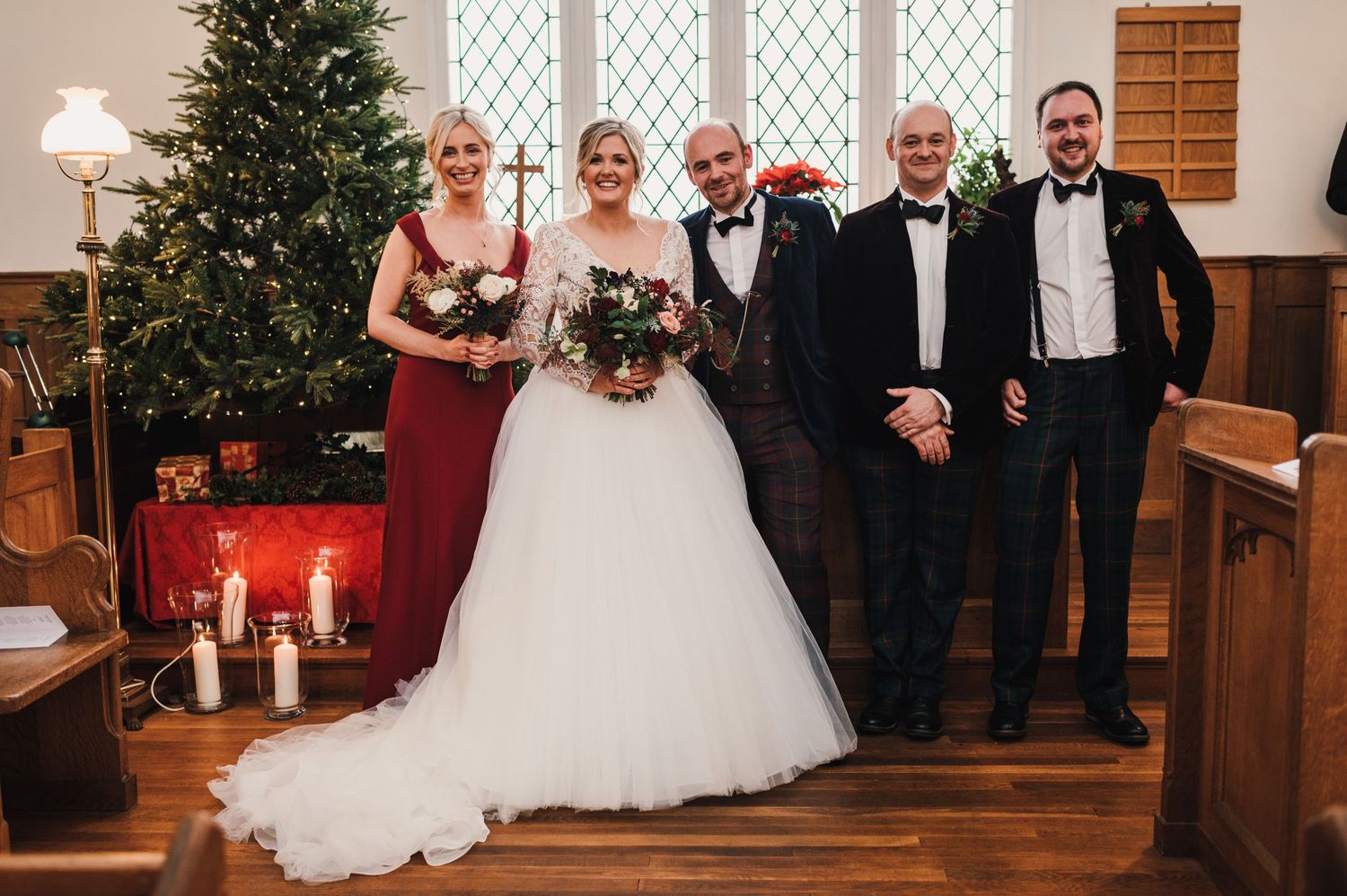 Wedding party poses together in church with decorated Christmas tree and wooden pews.