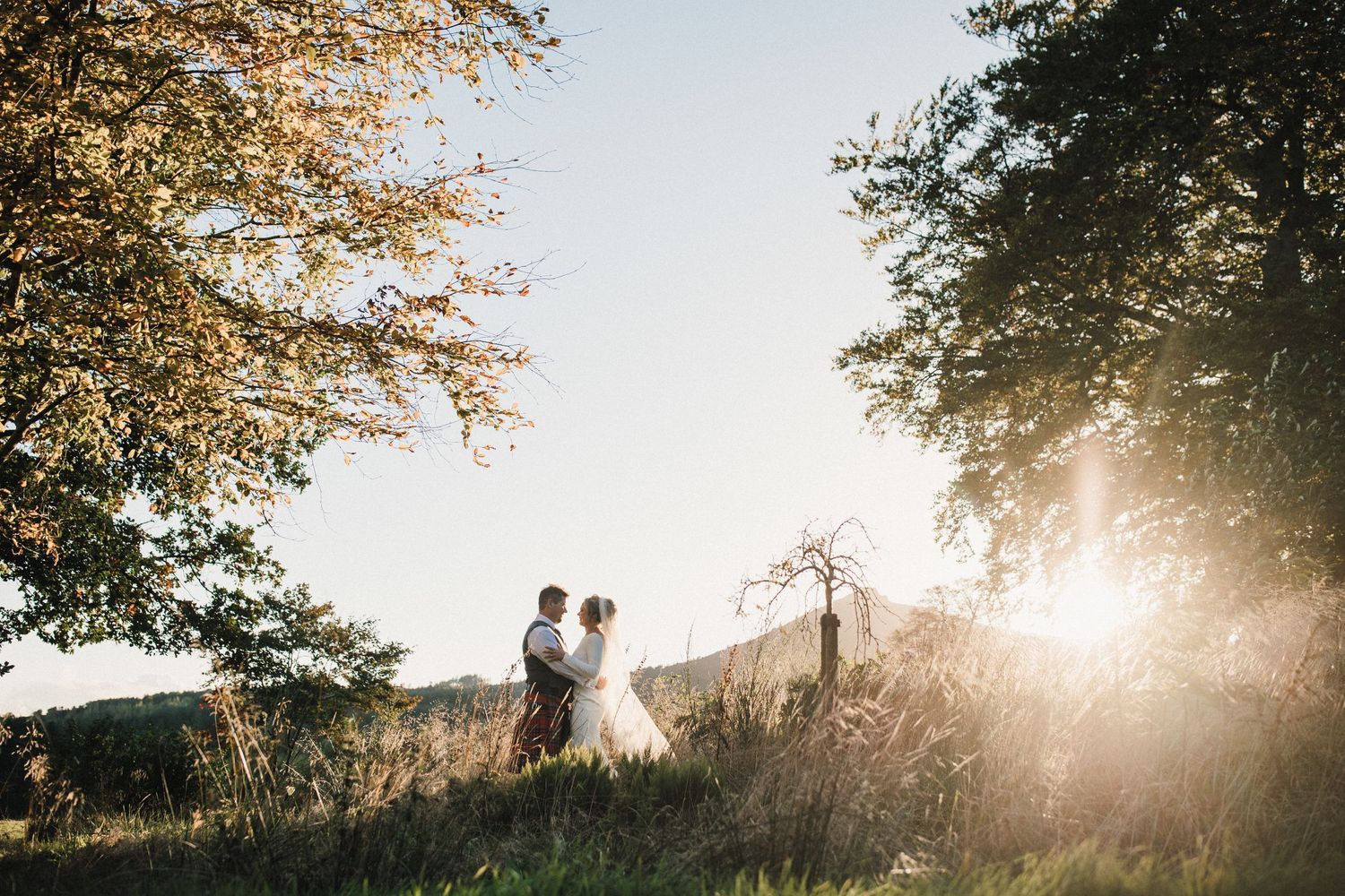 Silhouetted wedding couple standing on hilltop during golden hour with sun peeking through autumn trees.