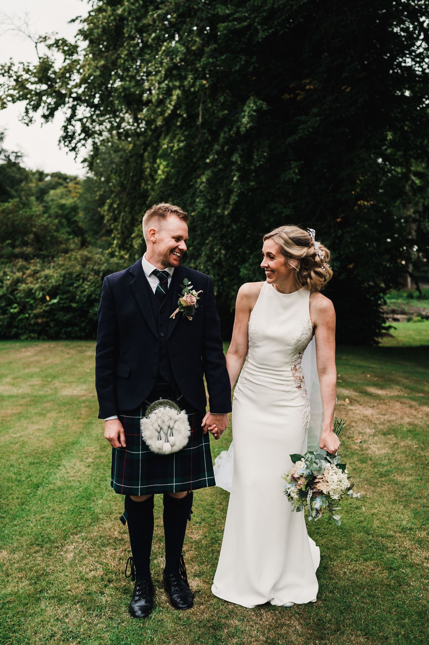 Elegant couple poses on lawn with groom in traditional Scottish kilt and bride in sleek white dress.