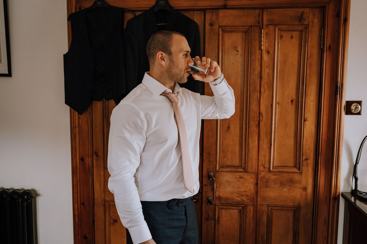 Side profile of person in white shirt fixing tie against wooden wardrobe.