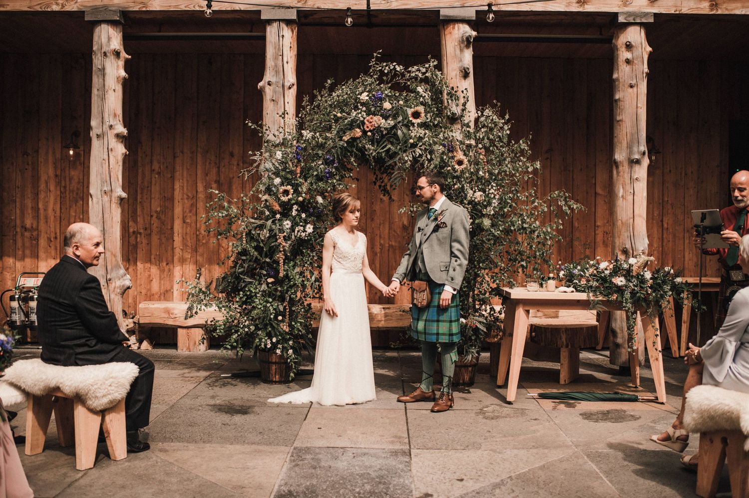Wedding ceremony under a floral arch with wooden backdrop and traditional Scottish attire.
