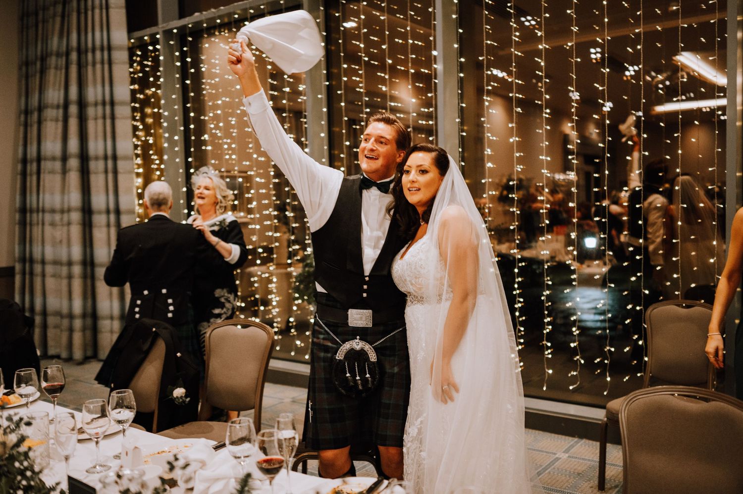 Couple celebrating at Scottish wedding reception with dramatic fairy light backdrop.