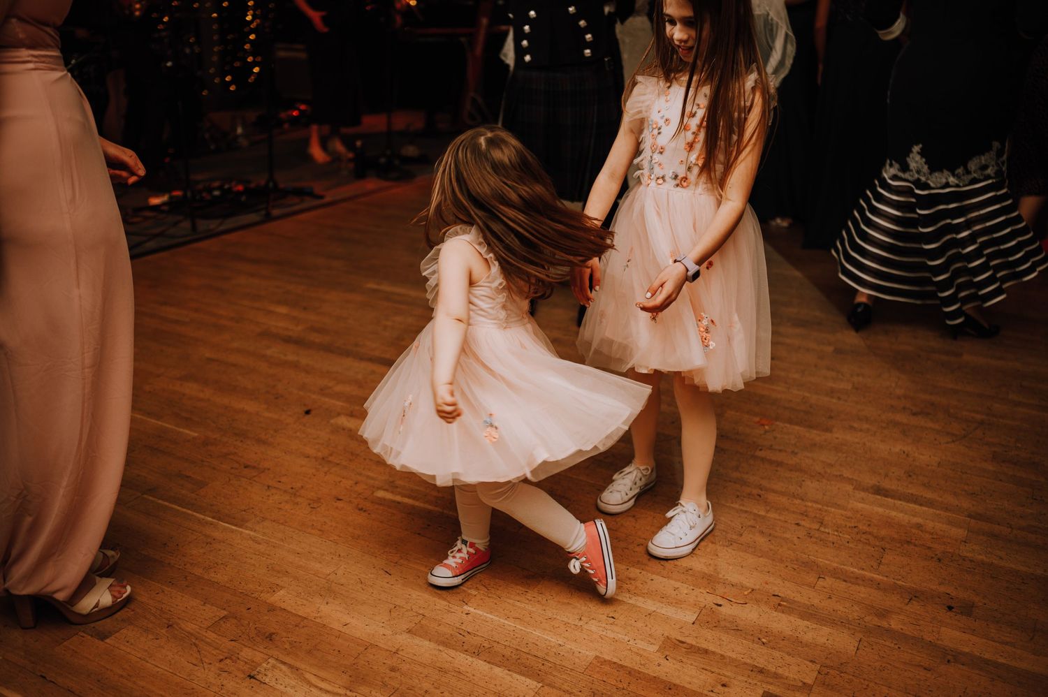 Two young attendees in pale pink dresses dance on a wooden floor at a wedding reception.