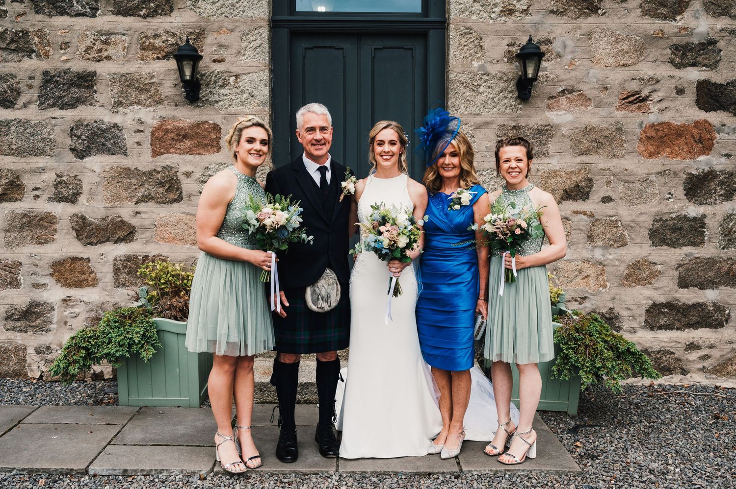 A wedding party poses outside a stone building wearing kilts and pastel dresses.