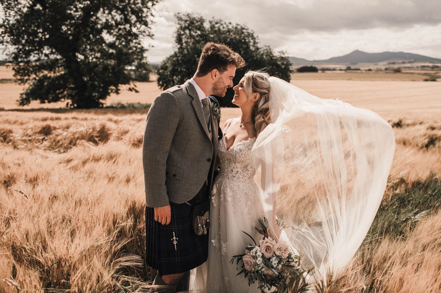 A Scottish wedding couple embraces in a field of wheat with dramatic cloudy skies.