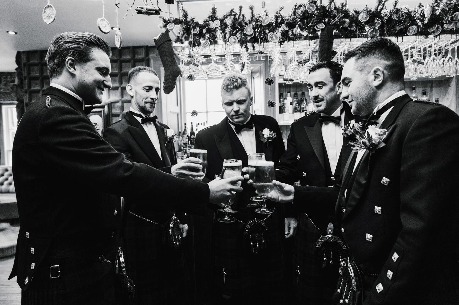 A black and white photo of groomsmen in formal attire toasting drinks at a celebration.
