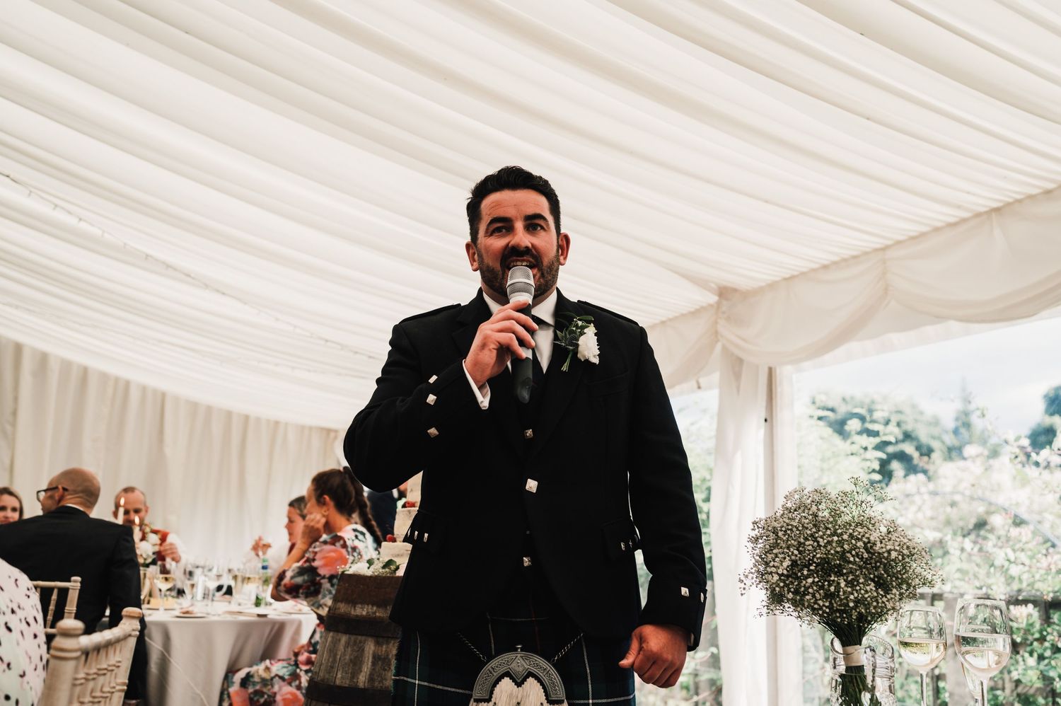 Speaker delivering a wedding speech in formal attire under white tent reception.