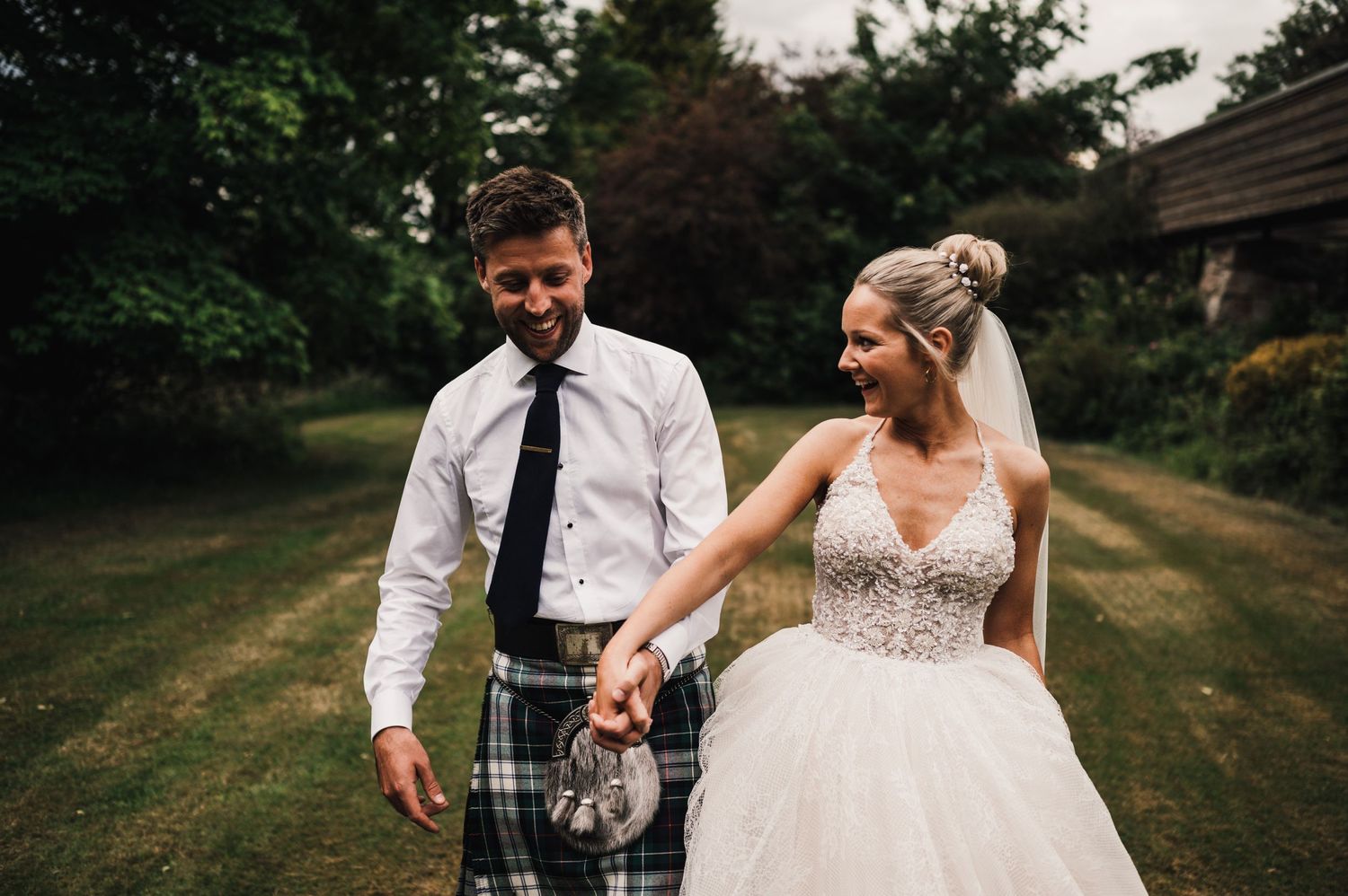 Candid wedding photo outdoors showing bride's elegant dress and groom's traditional Scottish kilt.